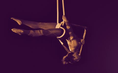 an aerialist hangs upside down, suspended by knotted rope against a dark backdrop