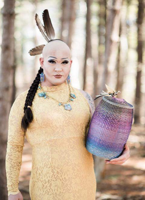 A person wearing a yellow dress and with Native American regalia and holding a multi-colored woven basket stands in a forest on a sunny day
