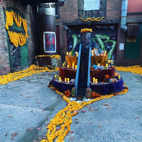 In an alley, an alter - with candles, yellow marigolds, sugar skulls, and other offerings - is shaped like a circular pyramid.