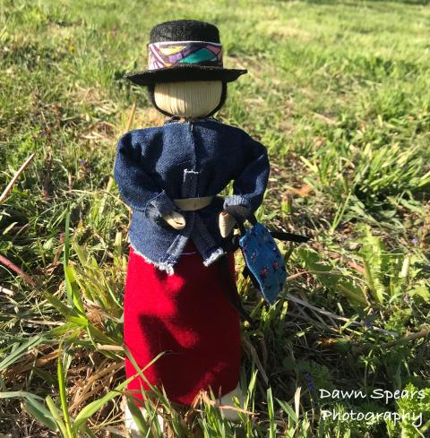 Corn husk doll dressed in black hat with colorful band, denim belted top, and long red skirt, standing on a sunny lawn.