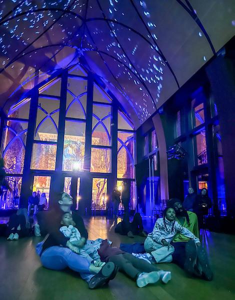 Folks sitting on the floor of a small chapel gazing up at projected illumination on the walls and ceiling