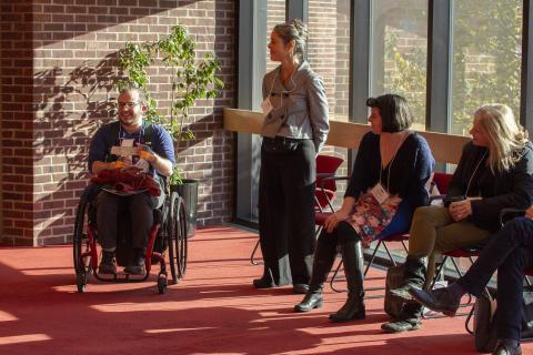  artist Toby MacNutt speaks to a group of people at the 2018 Idea Swap New to NEFA session. Toby is smiling and seated in their wheelchair next to other participants who watch them speak. Sunshine streams through tall glass windows behind the participants into the meeting room.