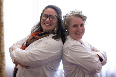 Two smiling, labcoat-wearing women pose back to back with their arms crossed 
