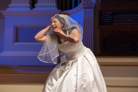 artist Sara Juli performs an excerpt of her new work (in development) titled "Burnt Out Wife" at the 2018 Idea Swap on the Great Hall stage at Mechanics Hall. She wears a wedding dress over her clothes. A wedding veil covers her face and a lanyard with a name tag hangs around her neck. Sara is slightly bent forward, holding the top of her head with her right hand and holding her left hand under her chin. She appears to cry with her eyes closed and mouth open. Blue stage lighting is seen behind her on stage.