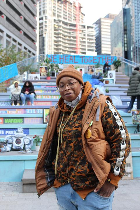 A Black woman, in brown winter gear, poses in front fo stairs with artwork on it.