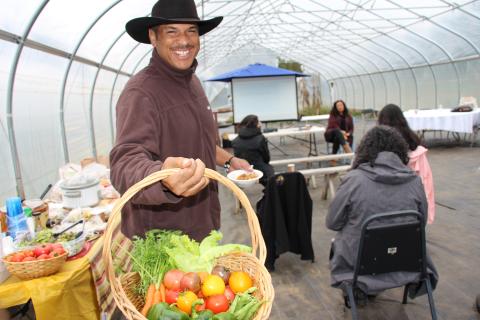 Cassius Spears, founder of the Narragansett Food Sovereignty Initiative and caretaker of the Crandall “minacommuck” Farm, smiles facing the camera while holding a basket full of  freshly picked carrots, tomatoes, green beans, and peppers in a high tunnel at the Markets & Marketing workshop. A lunch buffet table is behind Cassius and workshop participants are seated in the background. 