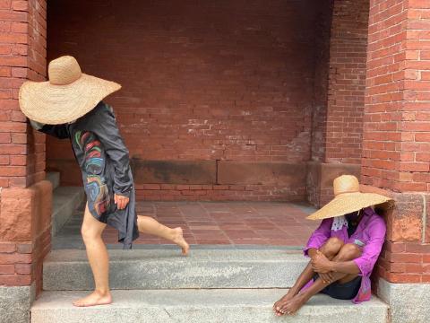 Two dancers, in sun hats, sit and dance in a brick entryway.