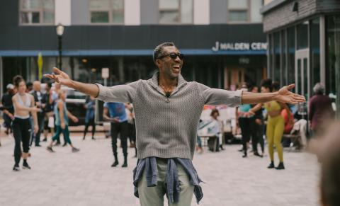 Outside, in a crowd of folks on pavement, a Black man, in sunglasses, holds his arms wide and smiles.