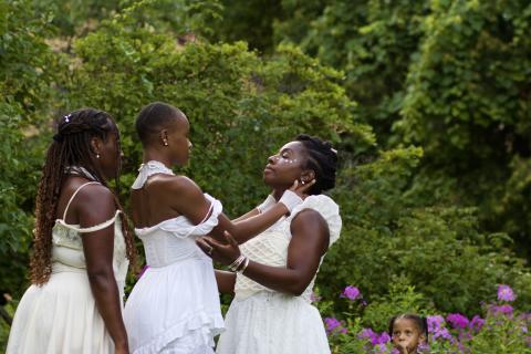 In a garden, three Black womxn in white gowns. One holds another around her face.