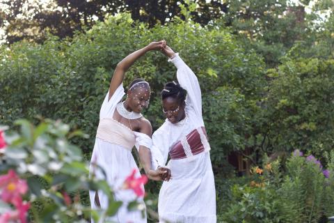 In a garden, two Black women, in white dresses spin with their hands clasped together and their backs to each other.