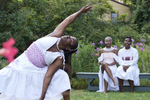 In a garden, a Black woman stands in a yoga pose, with her right arm to the sky and her left by her ankle. Two Black women watch from behind on a bench. They're all in white gowns.