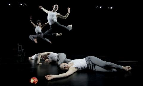 Two male dancers leap behind three female dancers who are unconscious next to an apple with a bite taken out of it.