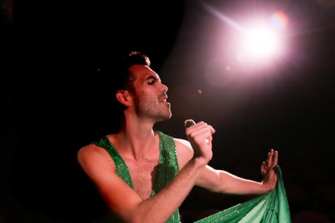 John sings into a microphone, while wearing makeup and a green dress, in front of a bright spotlight.
