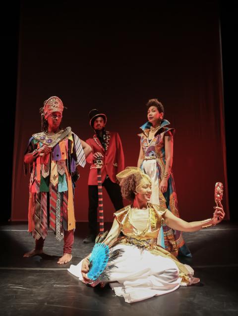 On a stage, four artists in regalia. Crowns, gold gowns, red blazers, dramatic collars, a turquoise fan. 