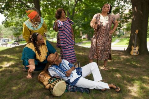 Five black women in traditional African dress pose with instruments under lush green trees