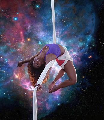 Pamela, a black woman in a unitard dangles from an aerial silk in front of a projection of the cosmos