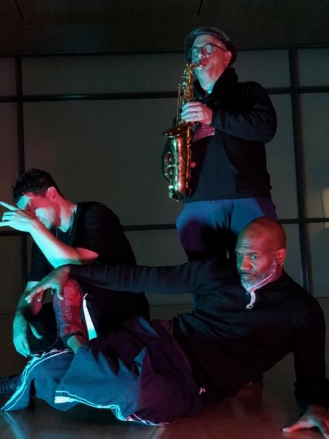 On a dimly lit stage, Xavier and a second dancer pose, in front of a saxophonist.
