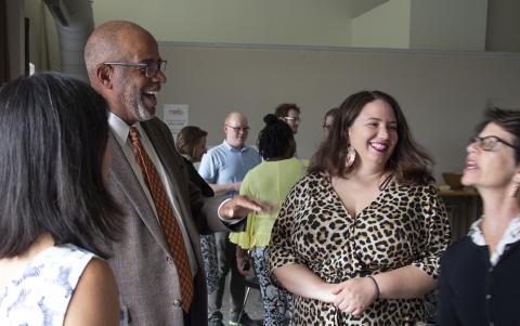 Four people conversing in a group, smiling and laughing. More people are standing and chatting in the background.