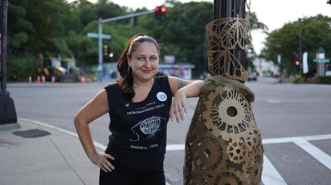 Ruth has light skin and long brown hair with red highlights. She leans on a pole with one a sculpture of gold gears wrapped around it.