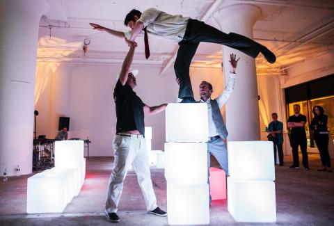 Two men help a woman who is standing on top of three light up boxes as she lifts a leg up behind her.