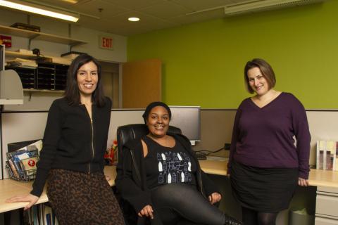 Three people pose and smile in an office cubicle.