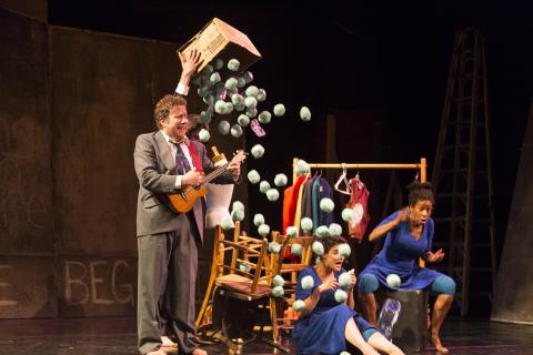 On a stage, a performer drops fake hail balls over a ukulele player and two performers wearing blue dresses.