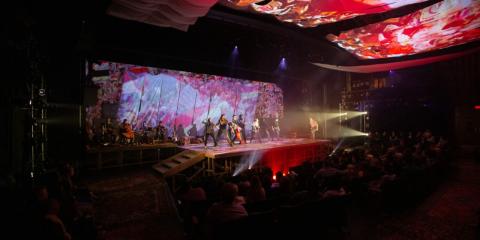 A dark theater with 8 actors facing the audience and a band in the background. Multicolored fabric and projections adorn the backdrop and the ceiling.