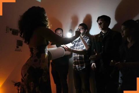 In a dark gallery space, Yara reads from a text, while an audience member holds a flashlight over the document.