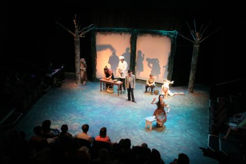 Young actors on a forest-decorated stage are dressed as fairy tale characters in front of a shadow figure scrim of more fairy tale characters.