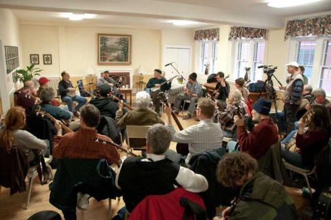 Inside a bright room, a group of thirty adults sit in a circle playing banjos.