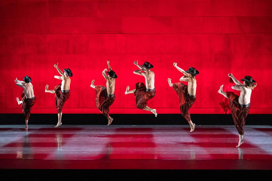 Men, in cowboy hats and red pants, leap in front of a red backdrop.