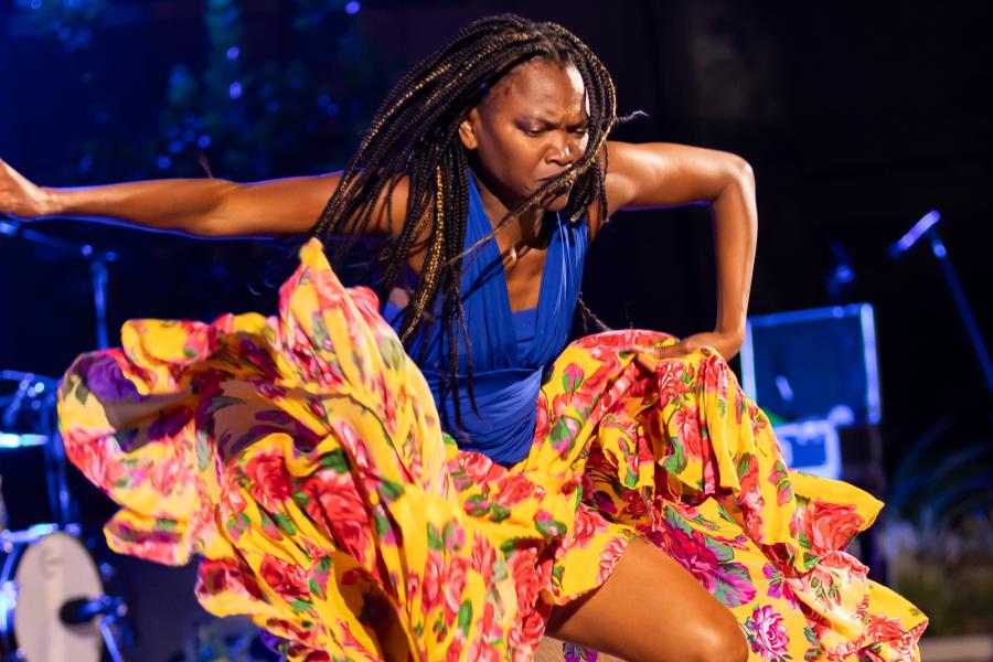 A Black woman, with long braids and a floral dress, leaps.