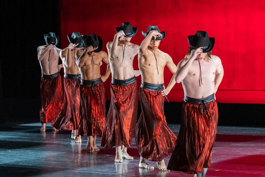 A line of male dancers, in red pants and in front of a red backdrop, hold their cowboy hats by the rim.