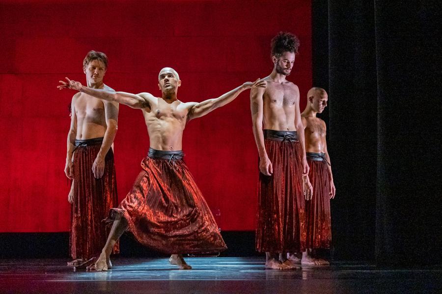 Four men, in red pants, perform on a stage in front of a red backdrop. One man holds his arms out and the others look down.