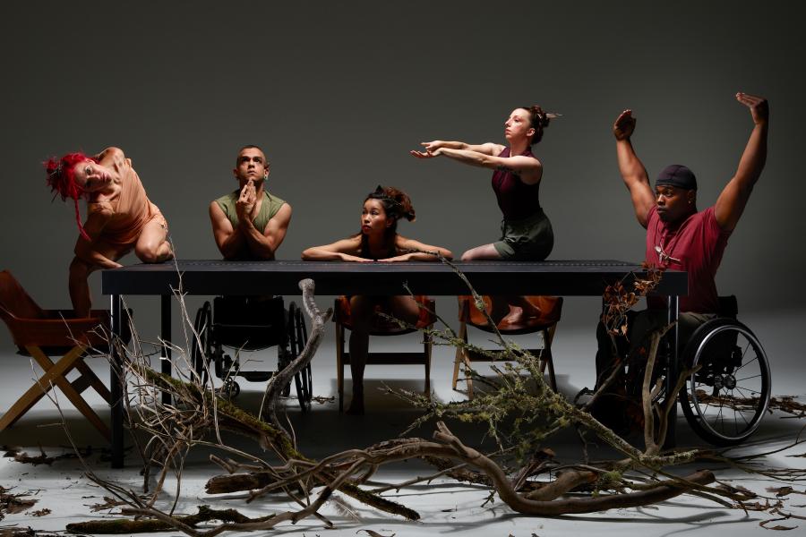 Five performers sit at or on a table. Two are in wheelchairs. One prays. Two hold their arms up or out.