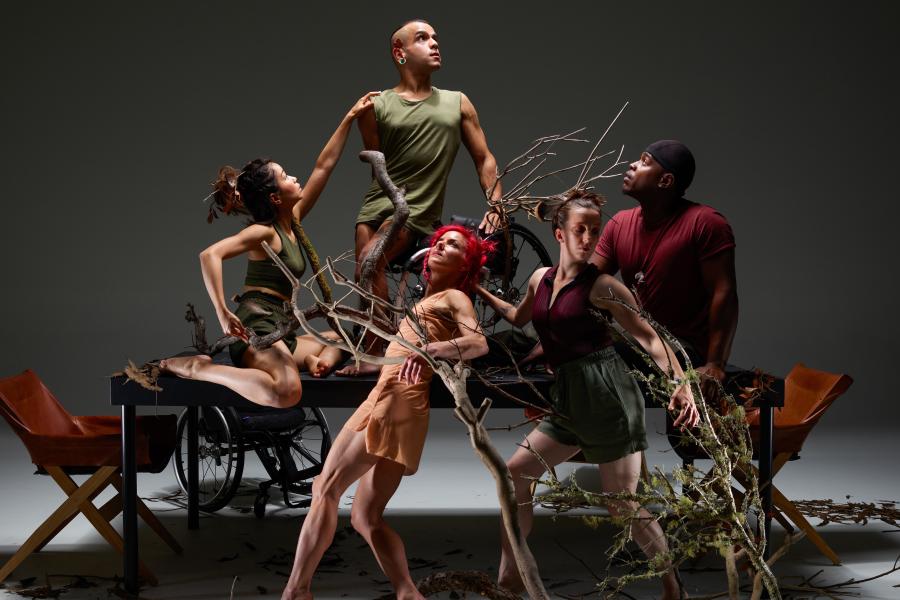 In front of a white backdrop, five performers climb a table and pose by shrubs  and large branches that have been placed on the ground.