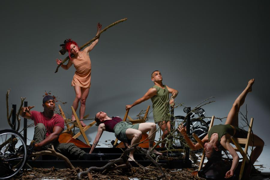 Five performers pose with branches and a couple lay across them, in front of a white backdrop.