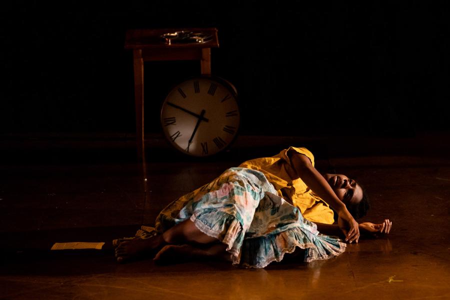 A Black woman lies on the ground in front of a very large grandfather clock.