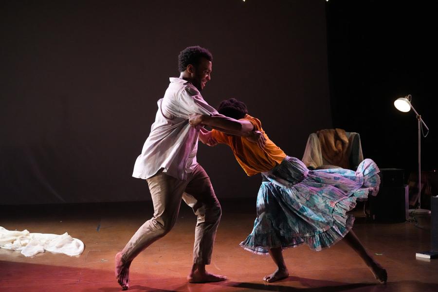 On a stage, with a chair and lamp, two Black folks dance. One of the dancer's dress spins out and away from her.