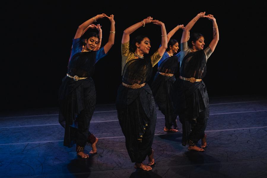 On a stage, a group of Indian women hold their hands up and dance in a circle in saris.