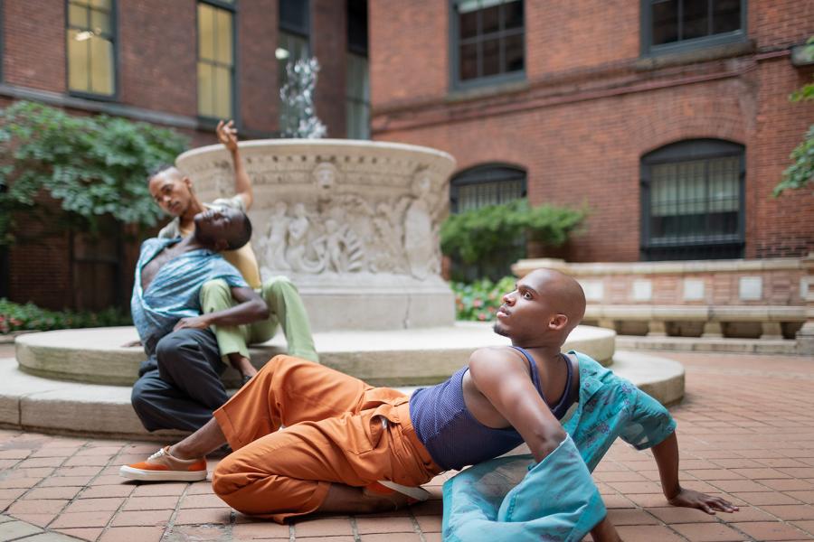 In a courtyard with a fountain, three dancers do floorwork in brightly colored casual wear.