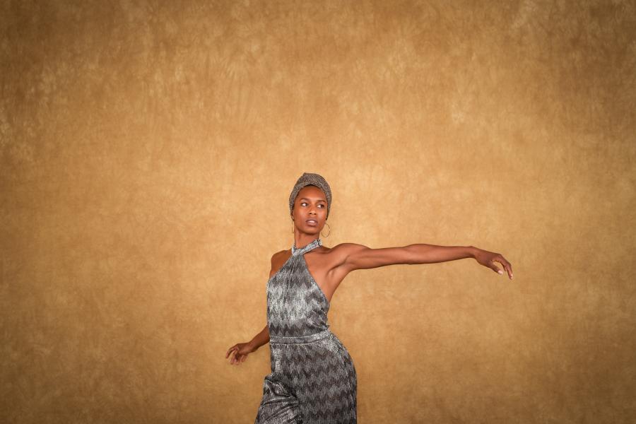 In front of a tan backdrop, a Black woman, in a grey headwrap and matching satin jumpsuit, holds her arm out to her left.