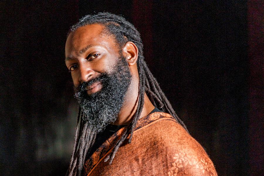 Antoine is a Black man with long braids and a long beard. A warm, rust colored fabric is draped over him.