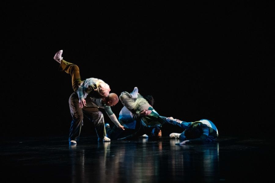 In a dark studio space with spotlights, five dancers perform connected by hand grabs and holds. One flops over the back of another.