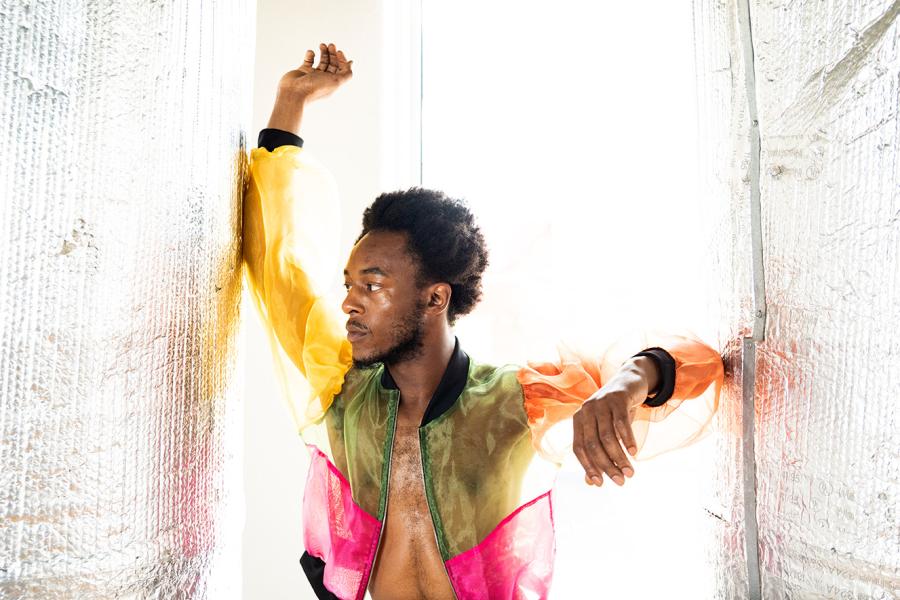 In bright light, a Black man wears a multicolored, sheer jacket and poses with his arms against walls on either side of him.