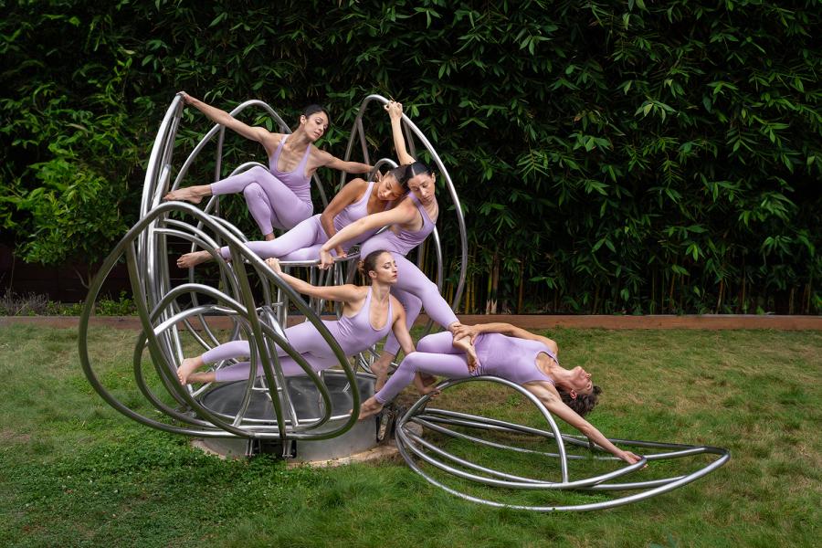 In a metal contraption that opens like a flower, one dancer leans over an open petal and four more lay inside of it.