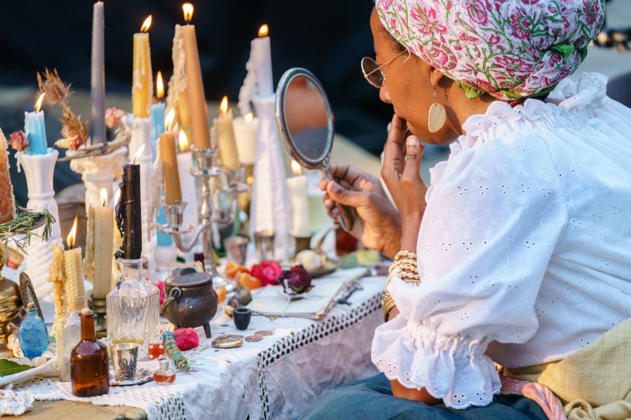 A brown woman looks into a mirror. She wears a headwrap. Behind the mirror is an altar with candles and offerings.