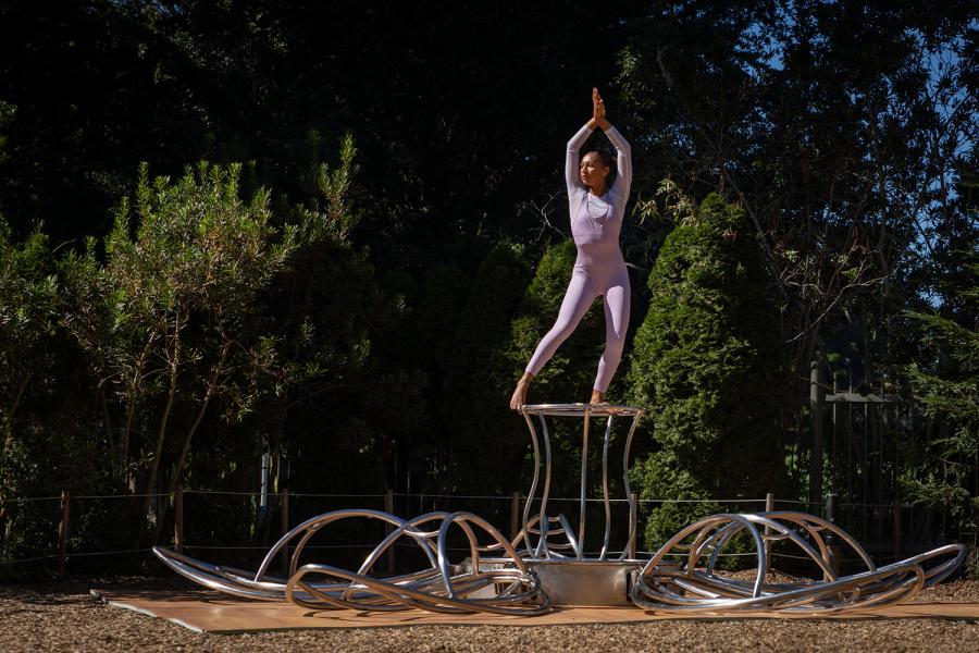 A woman, in a lilac full-body leotard, stands over a metal contraption that opens like a flower.