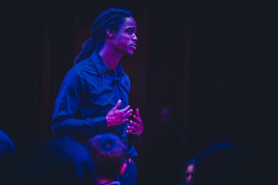 A Black man sings and holds his hands in front of himself. He is in a black box theatre. Blurred heads in the foreground.