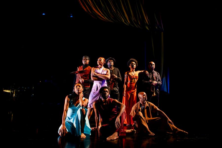 On a stage, a group of Black dancers pose in bright colors.